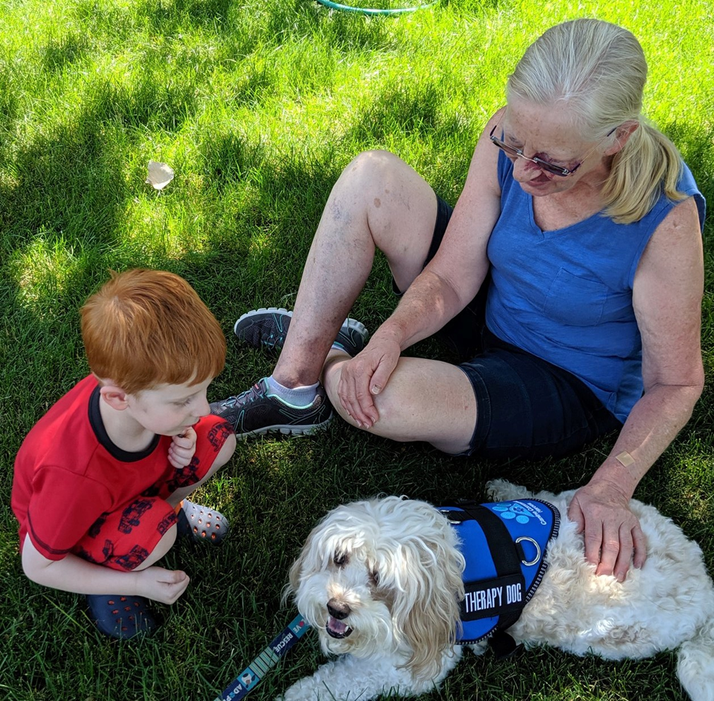 Woman and child sitting in the grass petting a small size dog therapy dog