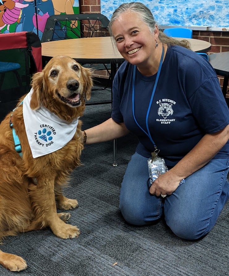 Jen VonLintel kneeling in a classroom with a school therapy dog sitting beside her